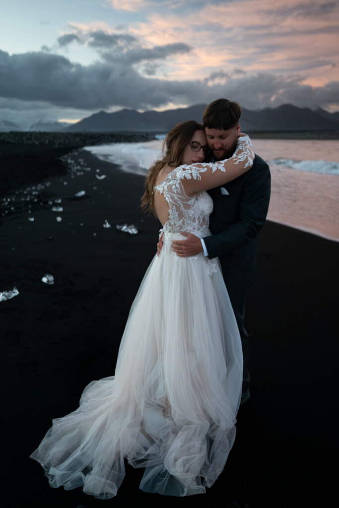 Couple embracing on an Icelandic black sand beach scattered with ice, beneath pink clouds at dusk.