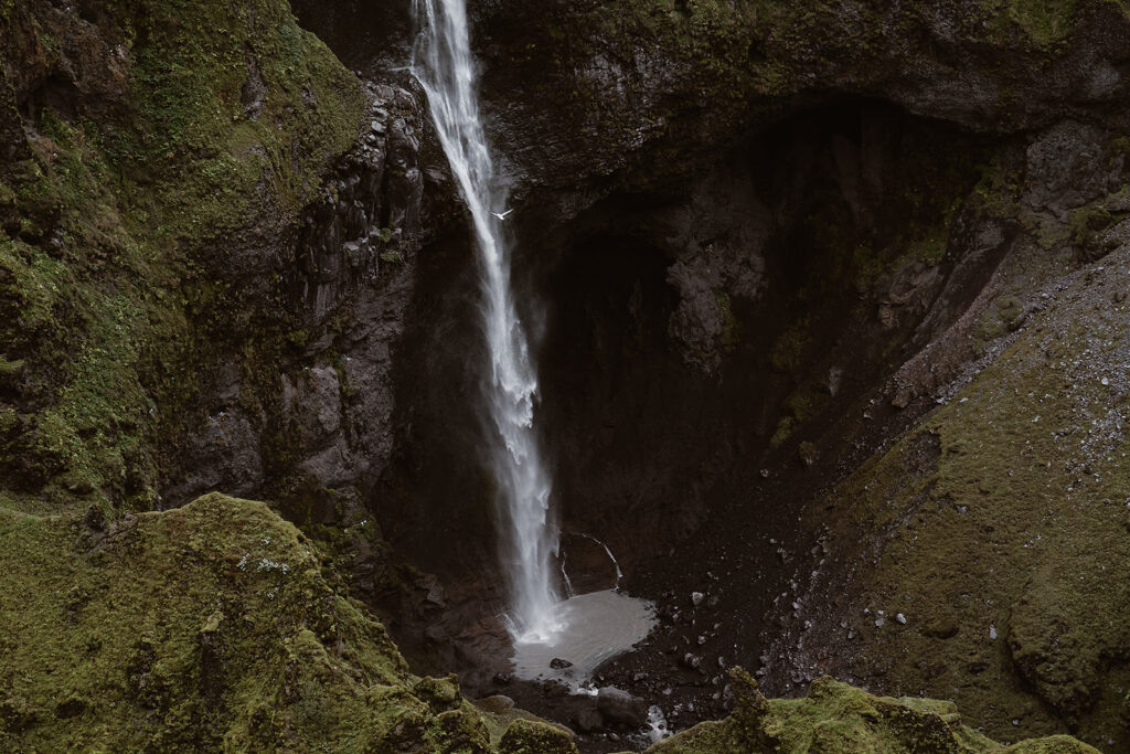 Tall, narrow waterfall plunging into a rocky pool between steep, moss-covered canyon walls in Iceland.