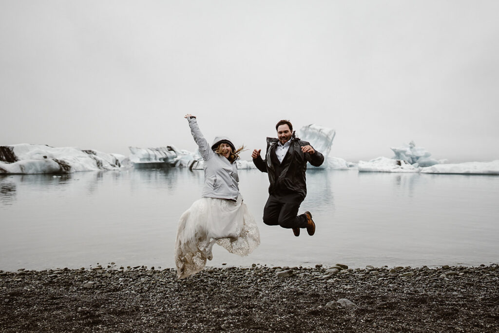 Couple in wedding attire and rain jackets jumping on a pebbled shore, with icebergs floating in an Icelandic glacier lagoon behind them at Diamond Beach.