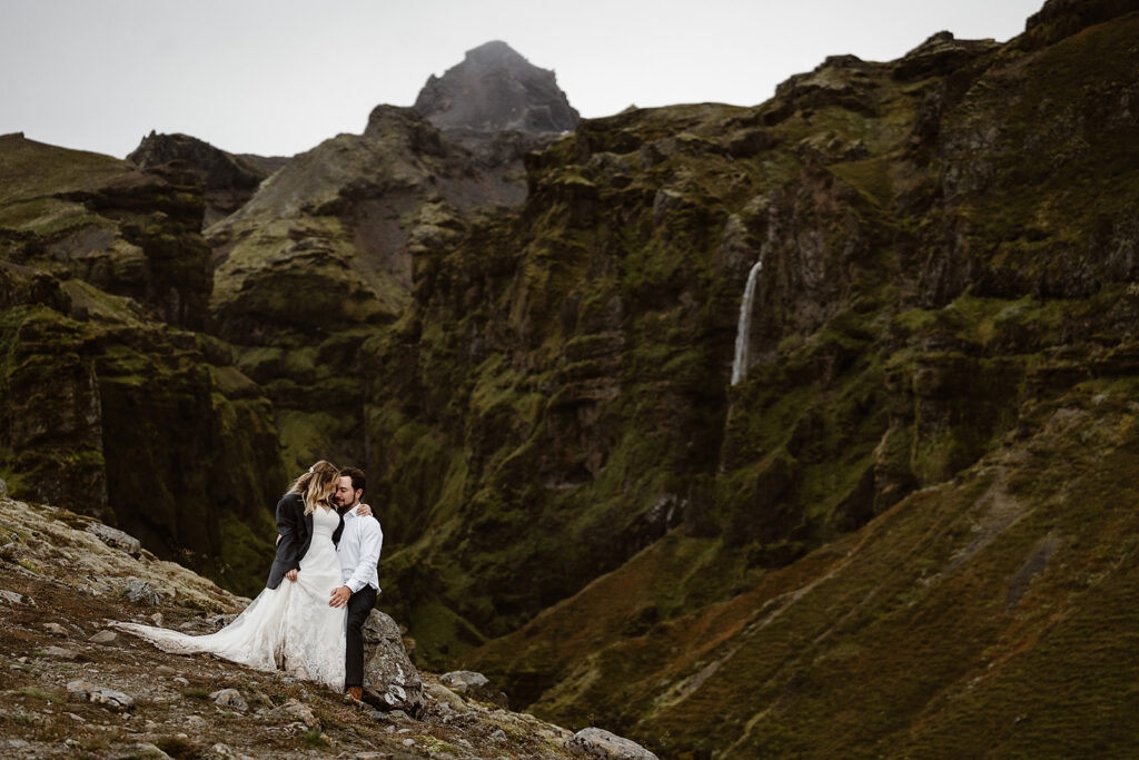 Couple embracing on a rocky slope above a moss-covered canyon in Iceland, with a narrow waterfall descending the cliff behind them.