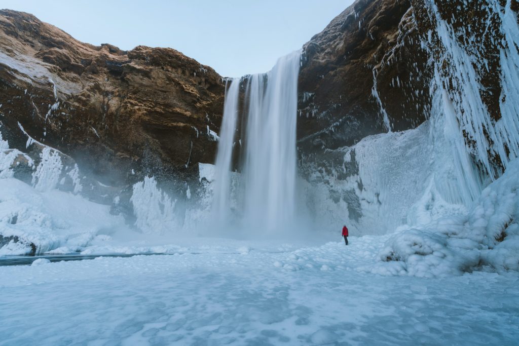 frozen over iceland waterfall with a small person in a red jacket standing under it