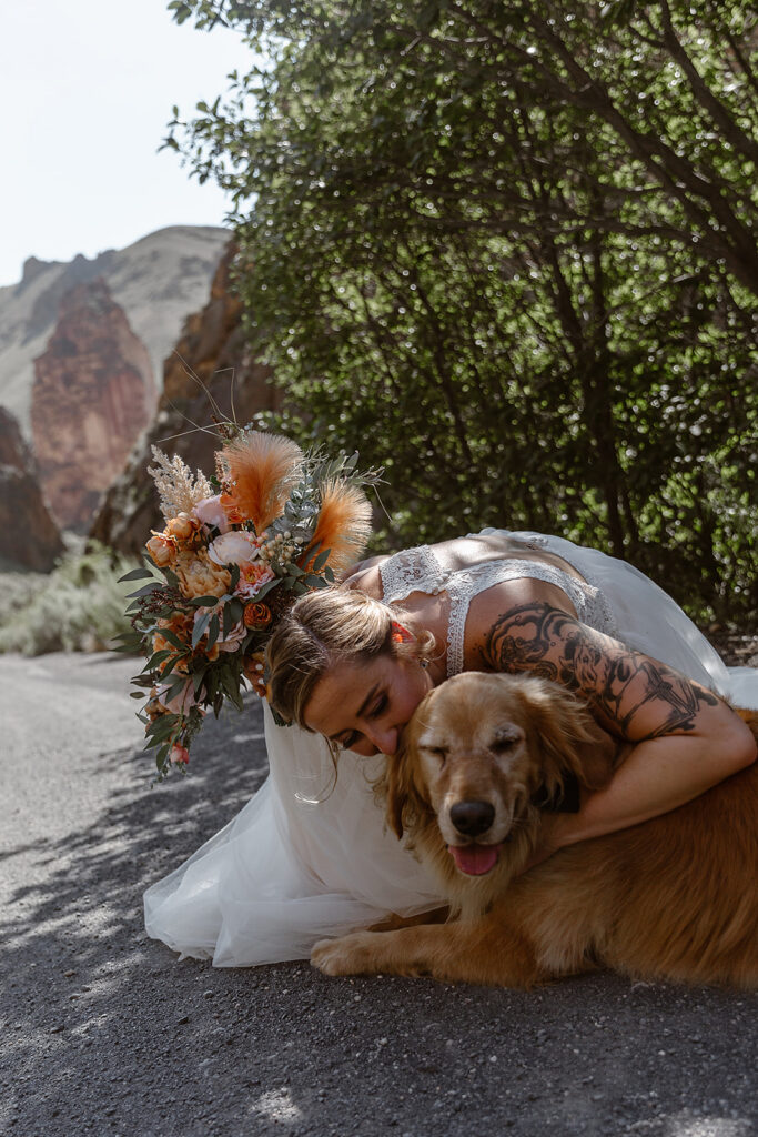 Bride hugging a golden retriever beside her colorful bouquet on a gravel road in Leslie Gulch.