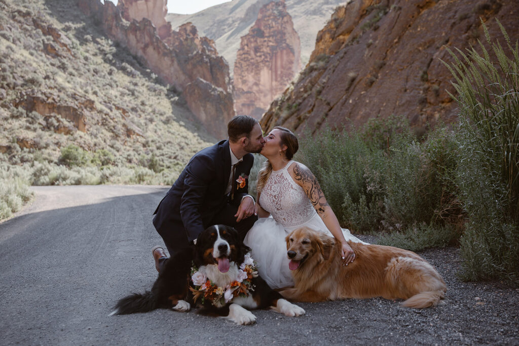 Newlyweds kissing while seated with their two dogs beneath Leslie Gulch’s orange rock formations.