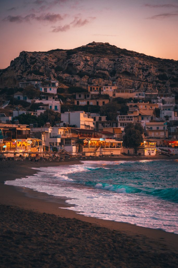 A wave rolls onto the shore with rows of stacked white houses in the backdrop during sunset in Crete, Greece