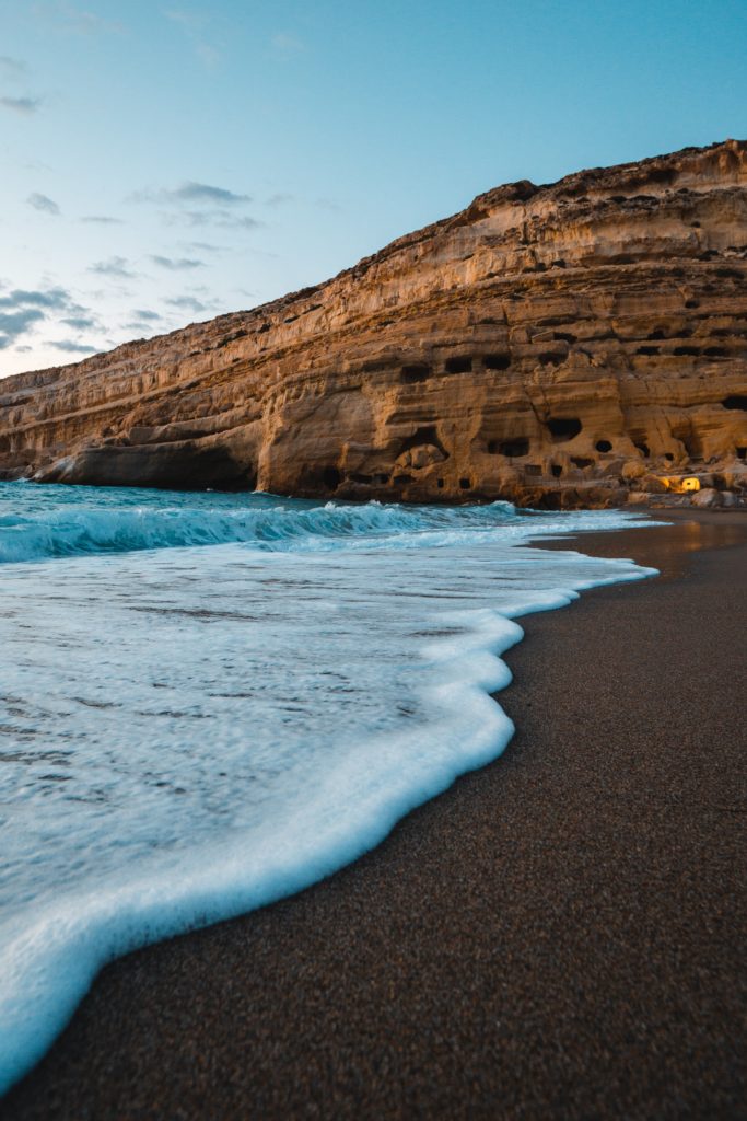 A wave rolls up on the shore of Matala Beach with a sandstone fixture in the background