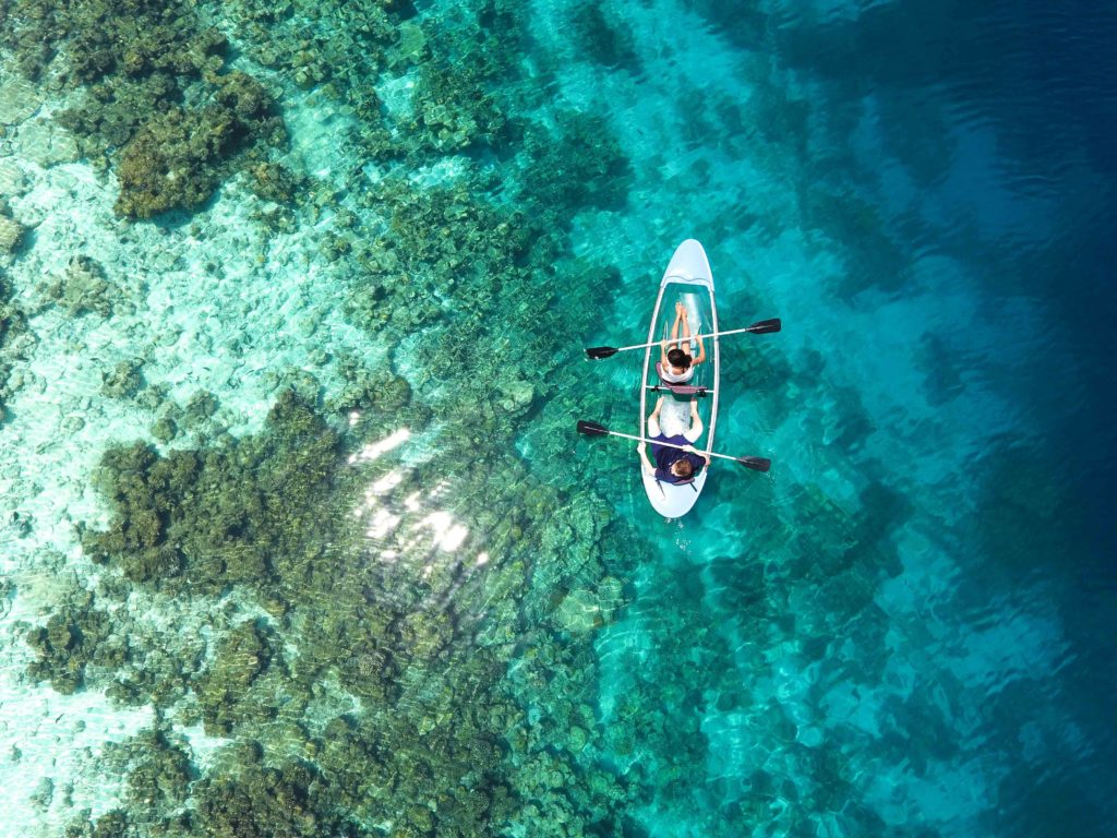 A couple paddles in a clear kayak above coral reefs in the Maldives