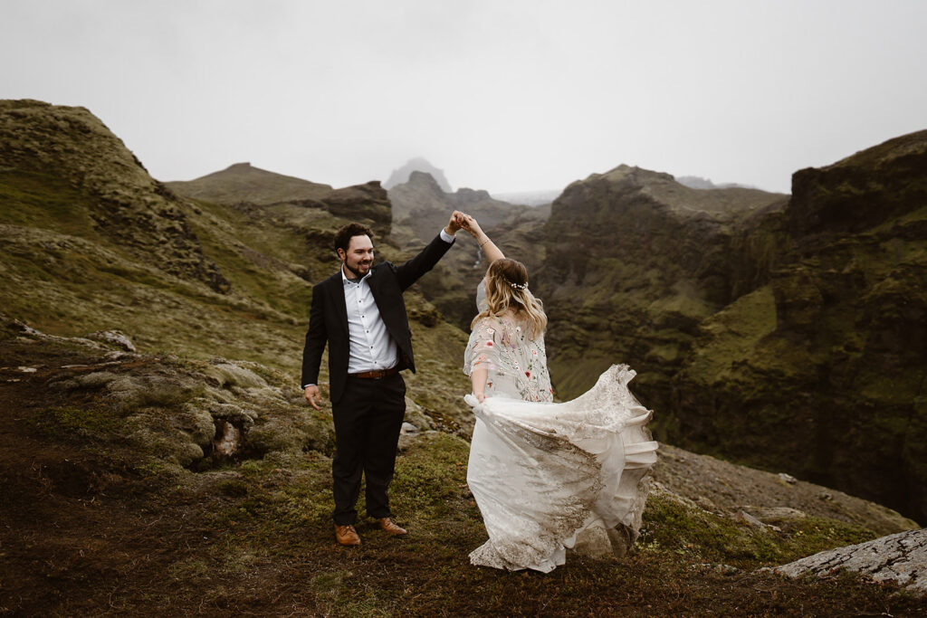 Groom twirling the bride on rocky ground in a moss-covered Icelandic canyon, her lace skirt lifting as she turns.