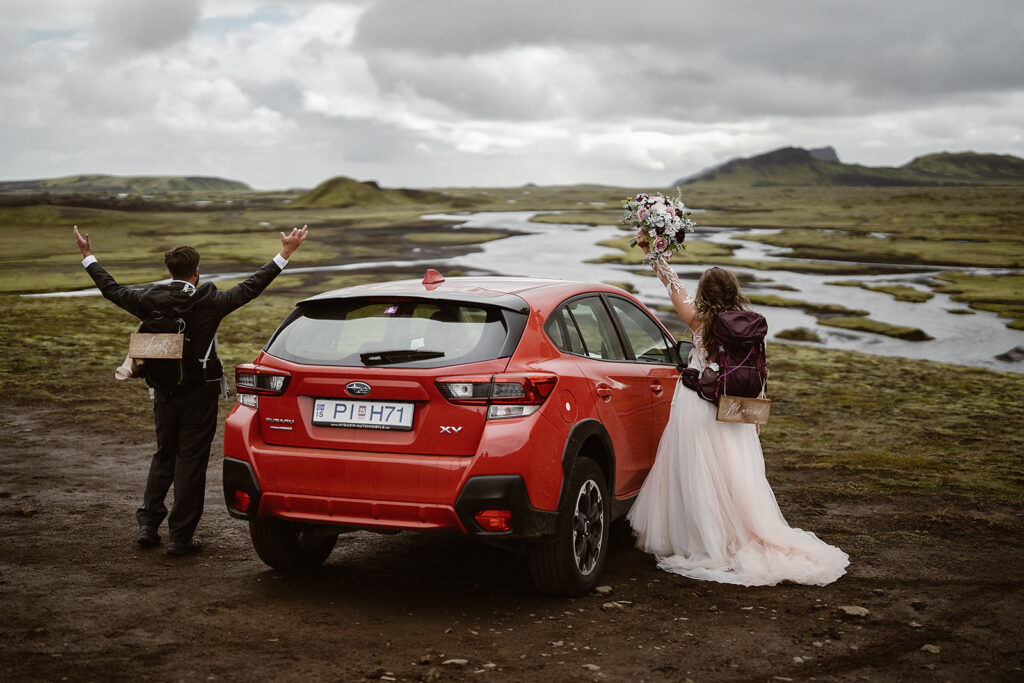 Couple wearing wedding attire and backpacks raising their arms beside a red car overlooking winding waterways in Iceland’s Highlands.