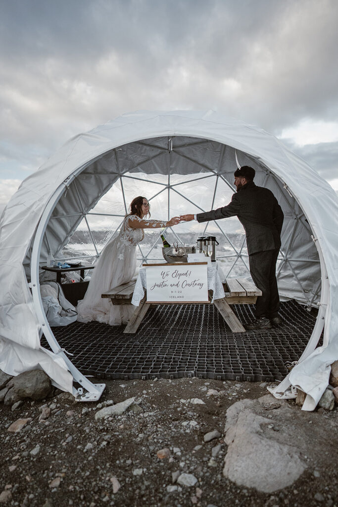 Newlyweds reaching toward each other across a picnic table inside an igloo, with a “We Eloped!” sign on the table.